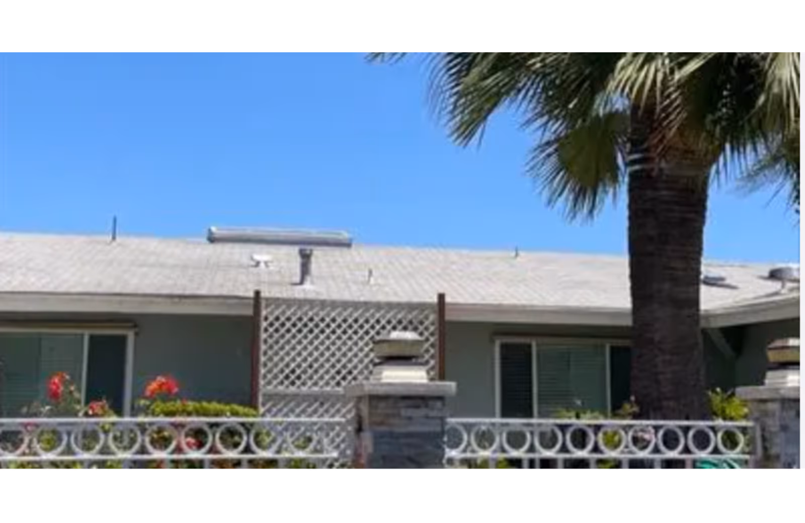 Front exterior of a single-story building with a white decorative fence, lattice panel, palm tree, and flowering shrubs under a clear blue sky.