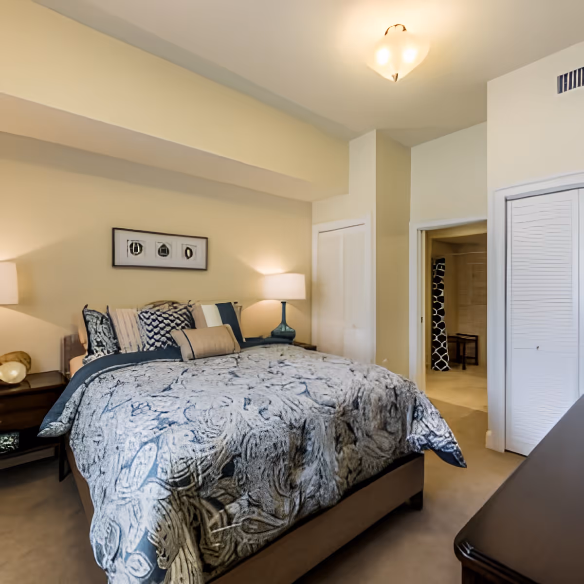 A cozy bedroom with a large bed covered in a patterned blue and white comforter, multiple pillows, two bedside tables with lamps, a framed artwork on the wall above the bed, a closet with white doors, and an open doorway leading to another room with a black and white patterned curtain.