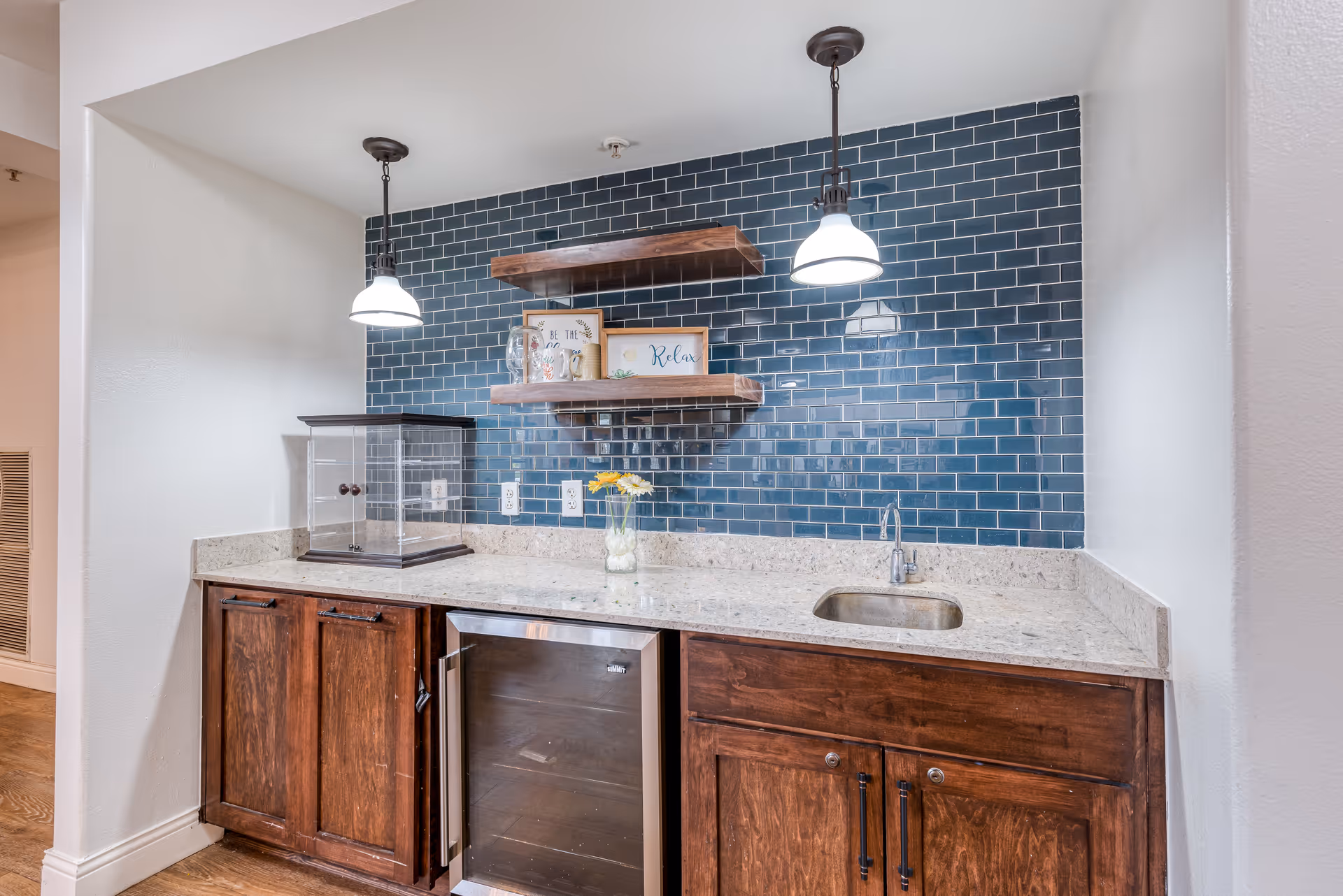 A kitchenette area with dark wooden cabinets, a small stainless steel sink, a mini refrigerator, and a countertop with a glass vase holding yellow flowers. The backsplash is made of glossy dark blue subway tiles, and two pendant lights hang from the ceiling. Two wooden floating shelves hold decorative items including framed signs and glass jars.