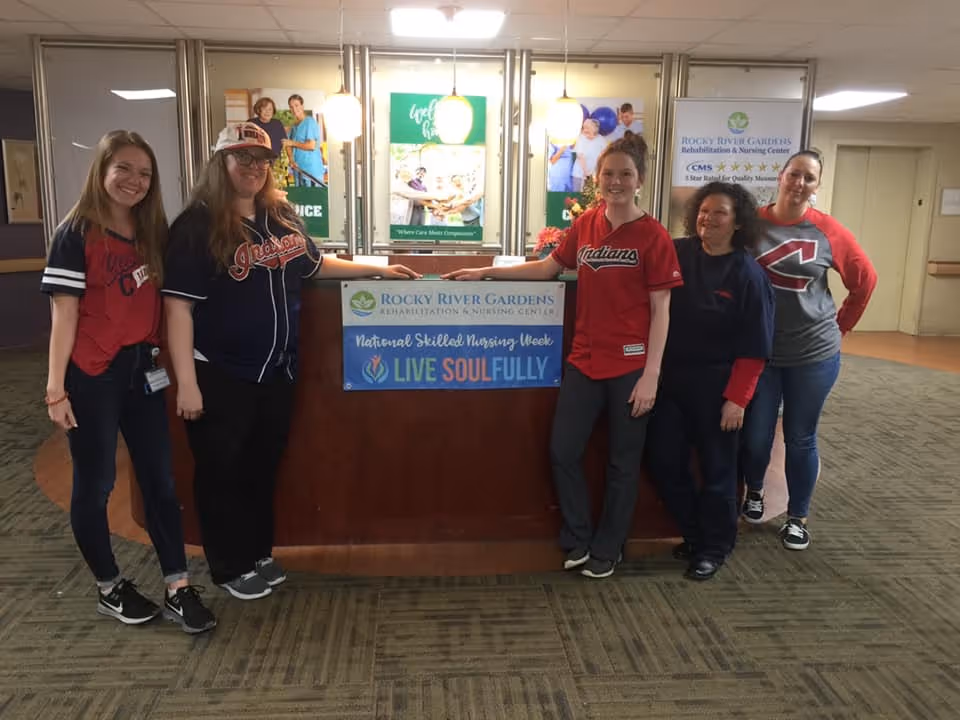 Five women standing inside a nursing center lobby behind a wooden reception desk with a sign that reads 'Rocky River Gardens Rehabilitation & Nursing Center National Skilled Nursing Week LIVE SOULFULLY'. The women are smiling and wearing casual clothing, some with sports-themed shirts. Behind them are posters and lights on the wall.