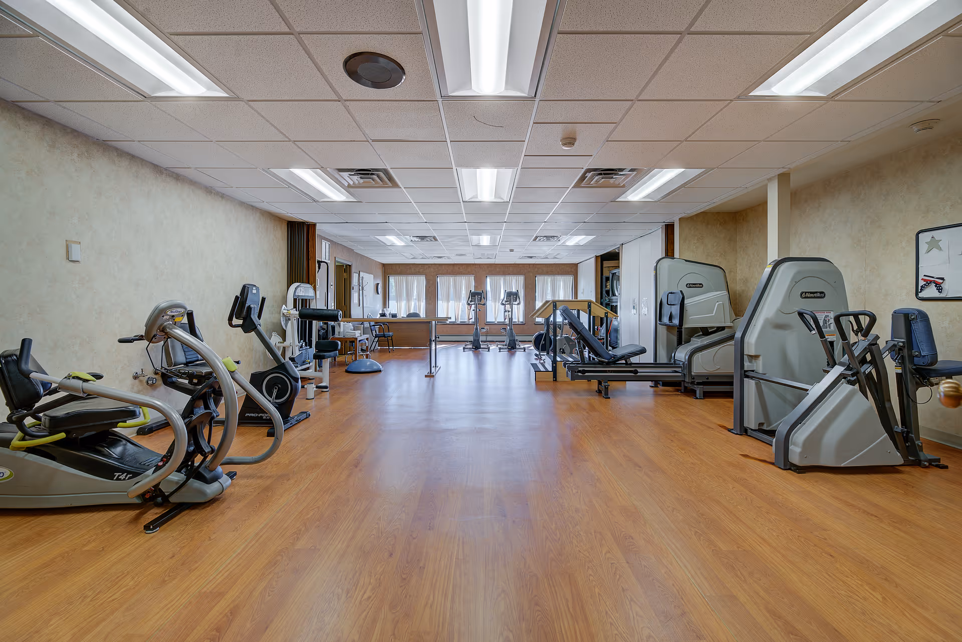 A spacious fitness room with various exercise equipment including stationary bikes, weight machines, and elliptical trainers. The room has wood flooring, beige walls, and a drop ceiling with fluorescent lighting. Large windows at the far end let in natural light.