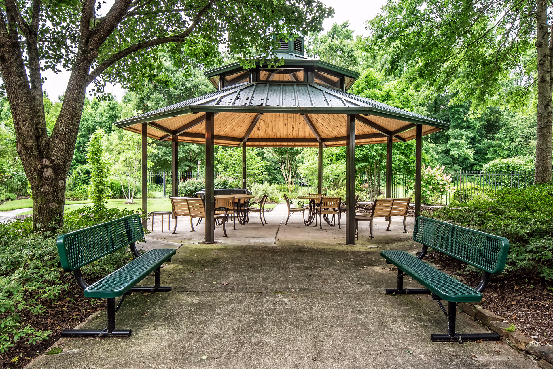 Outdoor gazebo with a metal roof and wooden ceiling, surrounded by green trees and bushes. There are several tables and chairs under the gazebo, and two green metal benches facing the gazebo on a concrete pathway.