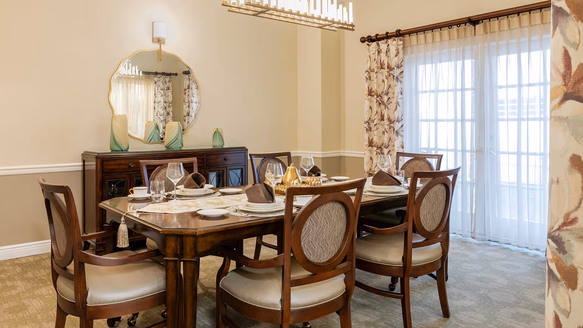 A well-lit dining room with a wooden dining table set for six people. The table has plates, glasses, napkins, and decorative items. There are six wooden chairs with cushioned seats around the table. A large mirror with a decorative frame hangs on the wall above a dark wooden sideboard. Sheer curtains and floral drapes cover a glass door leading outside.