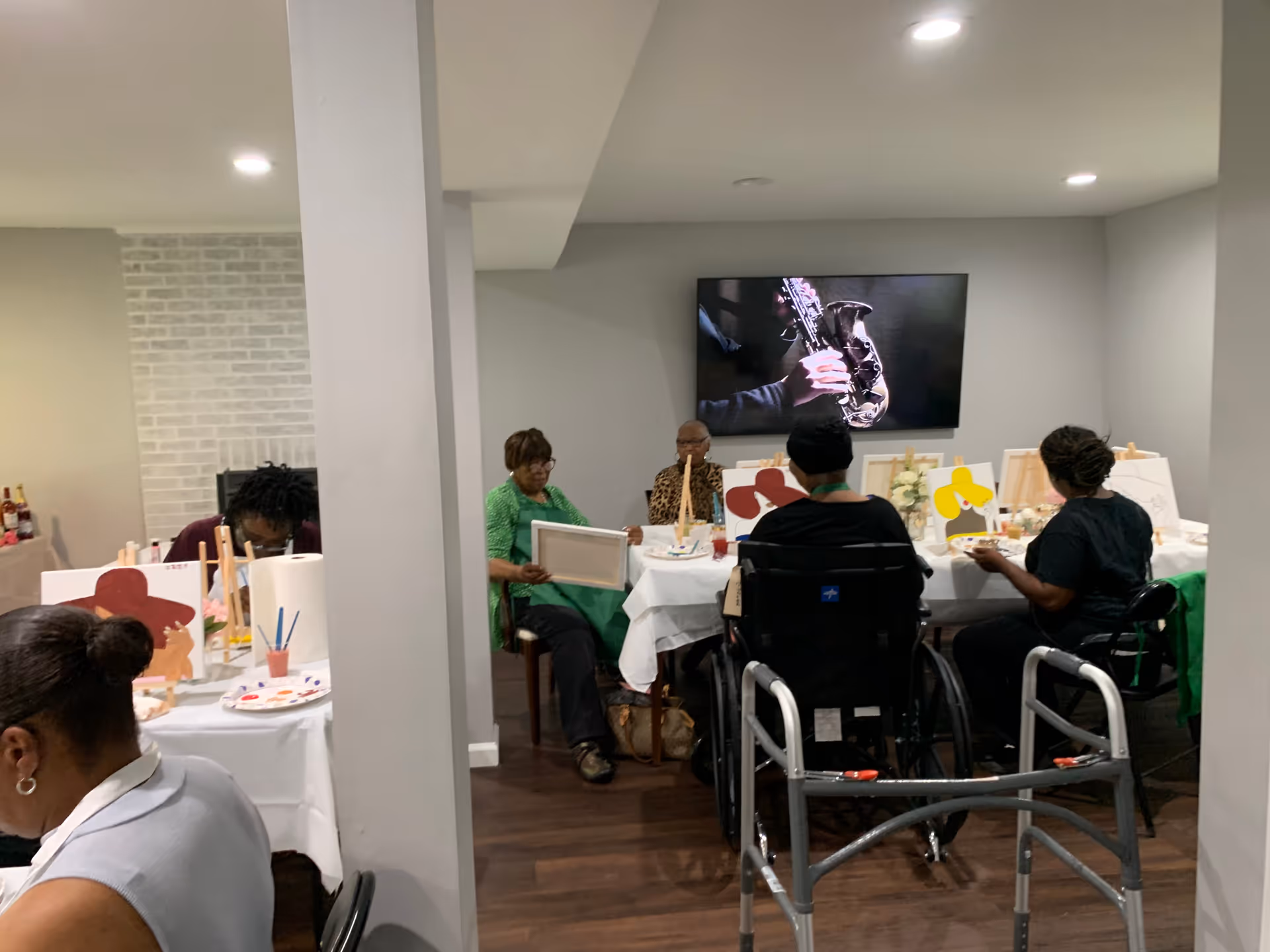 A group of elderly individuals seated around tables in a well-lit room, engaged in a painting activity with canvases and paint supplies. One person is in a wheelchair, and a walker is visible in the foreground. A large TV screen on the wall displays an image of a person playing a saxophone.