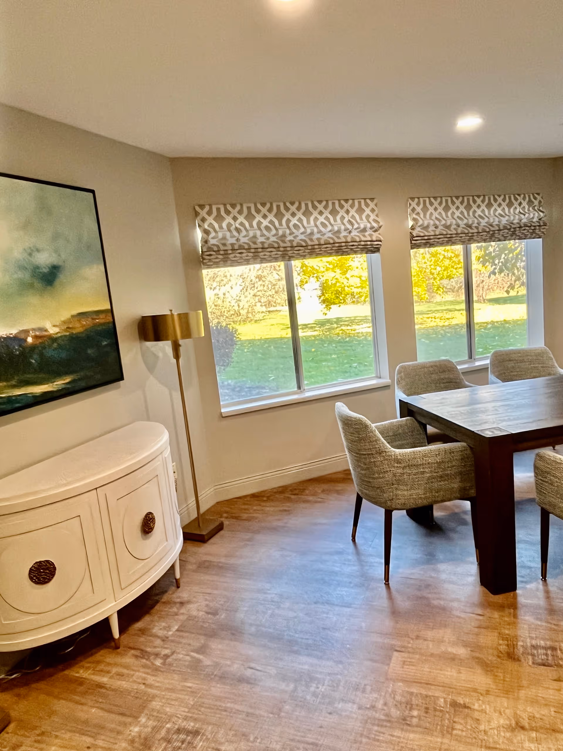 A bright dining area with a wooden table surrounded by four upholstered chairs. Two large windows with patterned Roman shades let in natural light and offer a view of green grass and trees outside. A white curved cabinet with decorative handles and a floor lamp with a gold shade are positioned against the wall, which also features a large abstract painting.