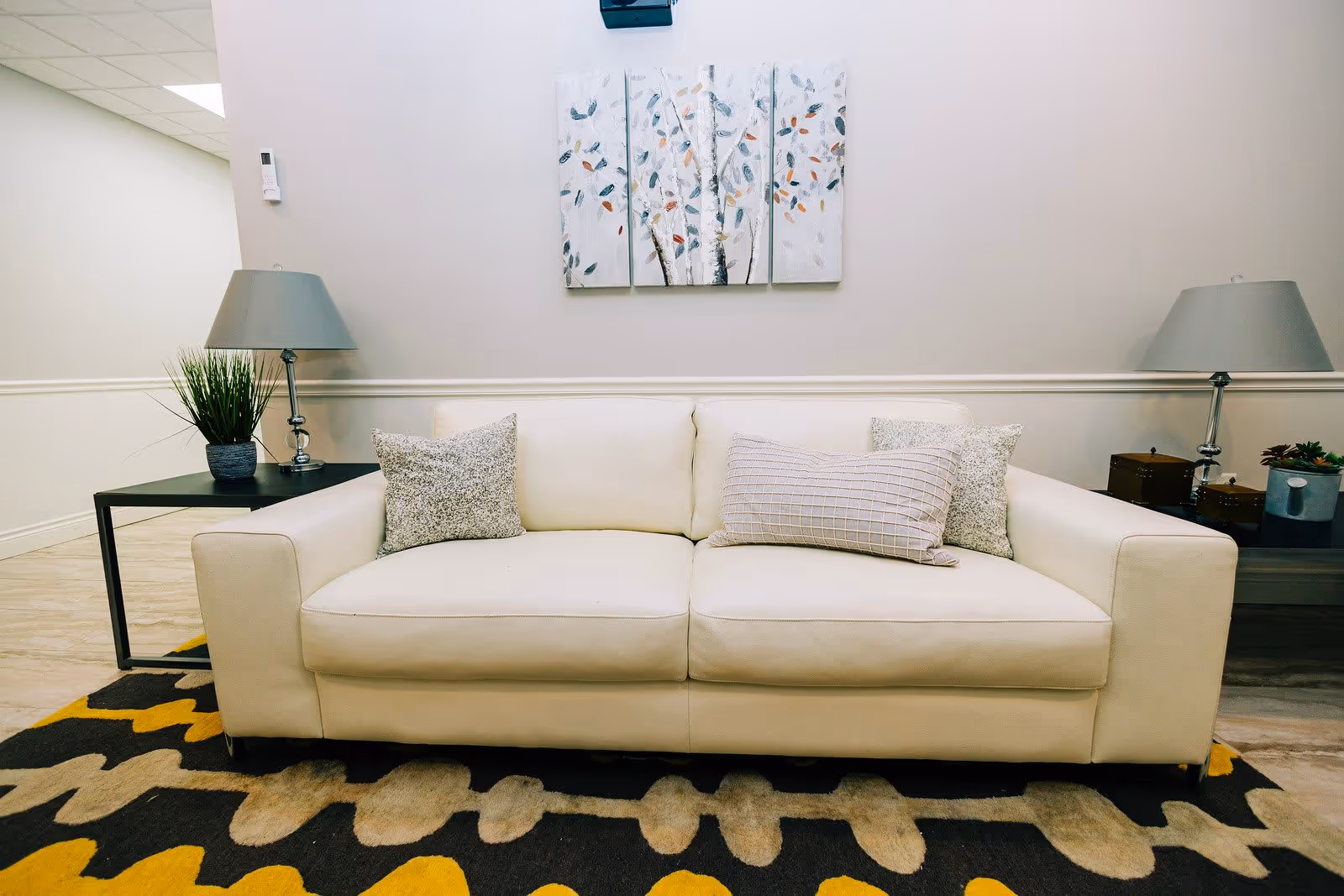 A white leather sofa with three decorative pillows is centered against a light-colored wall. On either side of the sofa are side tables with lamps and small plants. Above the sofa is a three-panel artwork featuring abstract tree designs with colorful leaves. The floor has a patterned rug with yellow, black, and beige colors.