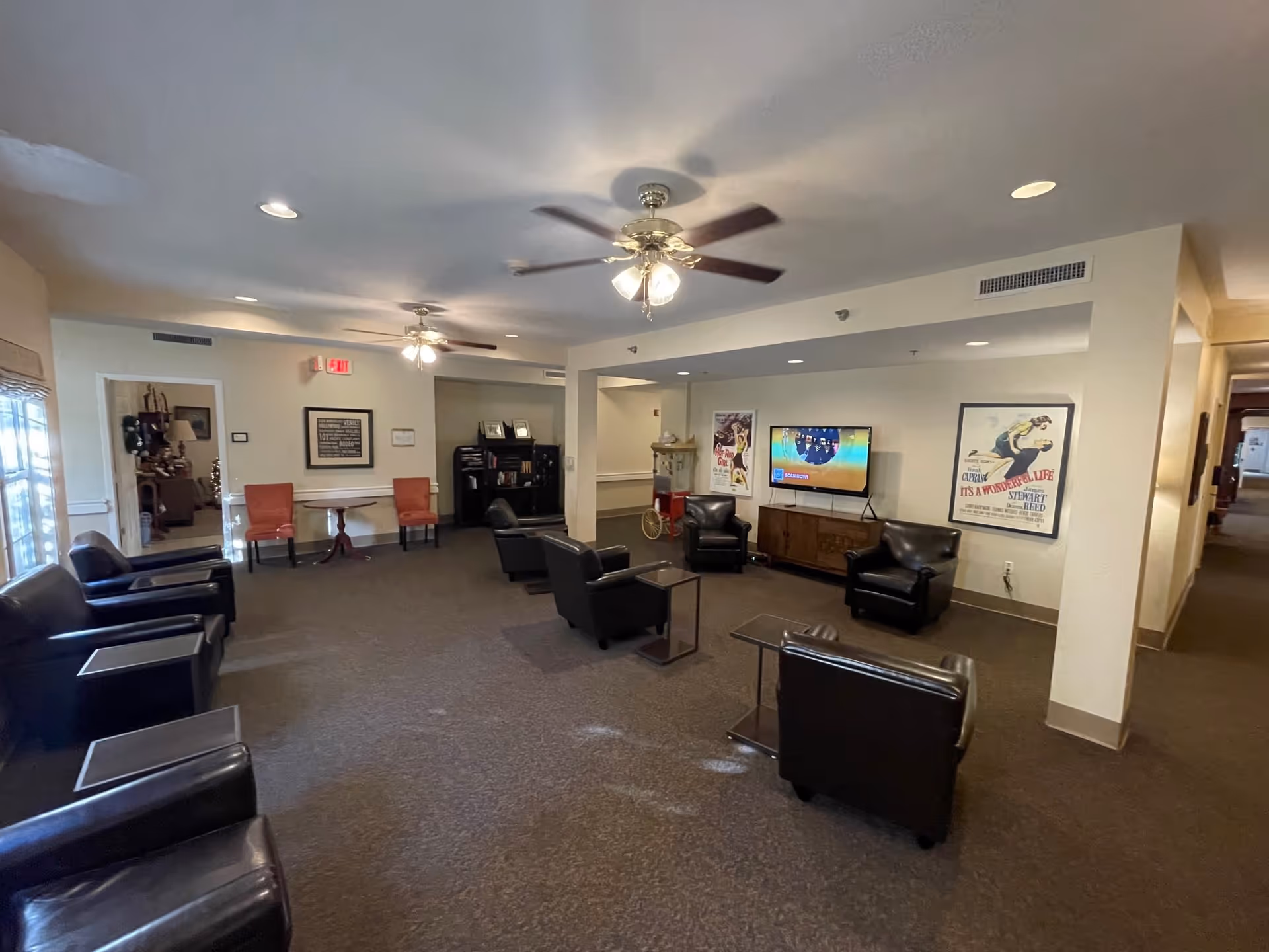 A spacious living room area in a senior living facility with multiple black leather armchairs arranged around small tables. The room has ceiling fans with lights, a flat-screen TV mounted on the wall, and framed vintage posters. There is a hallway visible to the right and a doorway leading to another room on the left.