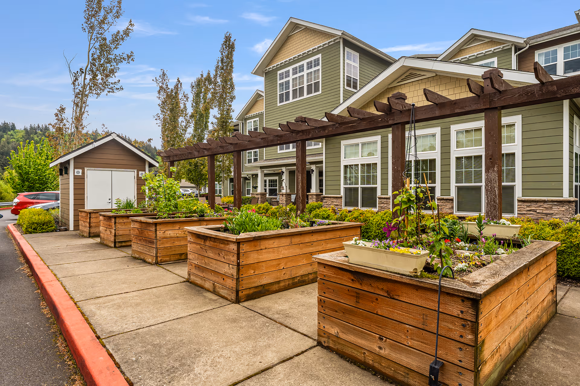Outdoor garden area at Glenmoore Gracious Retirement Living featuring raised wooden planter boxes with various plants and flowers, a wooden pergola structure, and a green multi-story building with white-trimmed windows in the background under a partly cloudy sky.