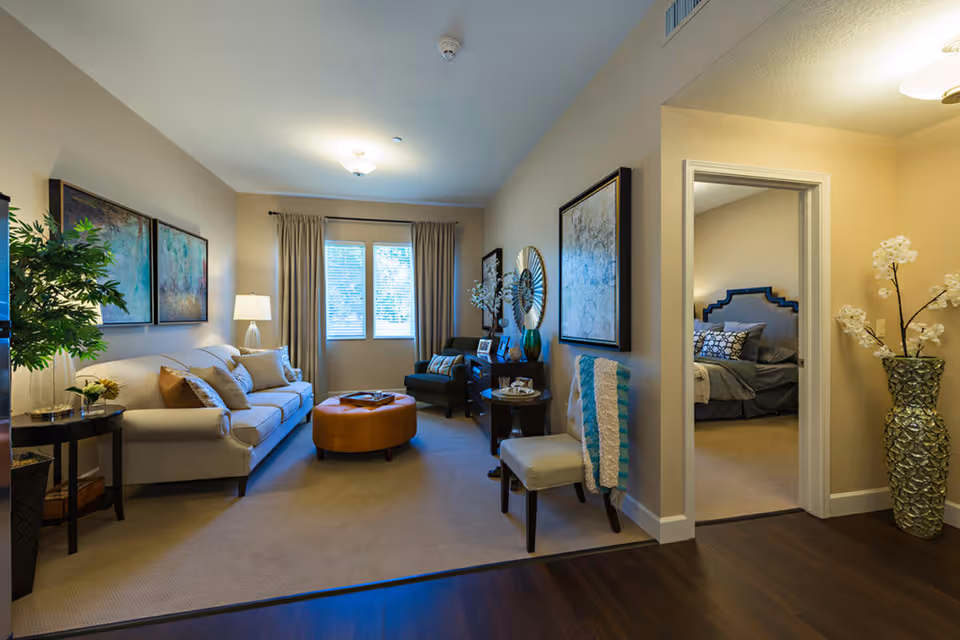 A cozy living room with a beige sofa adorned with pillows, a round orange ottoman, a dark armchair, and a side table with a lamp. The room has a window with curtains letting in natural light. Adjacent to the living room is a bedroom with a bed featuring a dark headboard and patterned pillows. The area has warm lighting, wall art, a chair with a throw blanket, and a tall decorative vase with white flowers near the entrance to the bedroom.