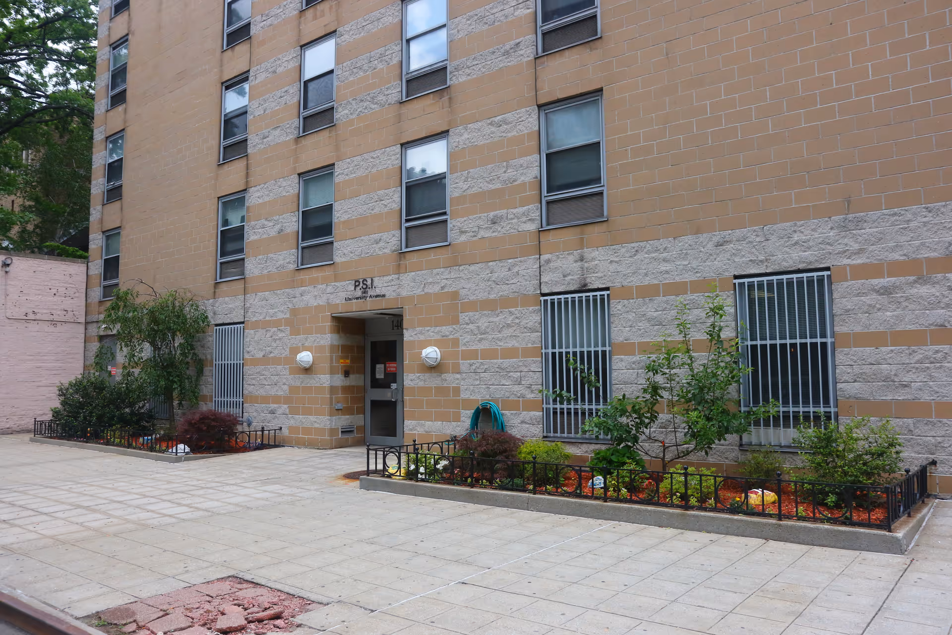 Entrance of a multi-story brick apartment building with small landscaped planters, barred windows, and a paved forecourt.