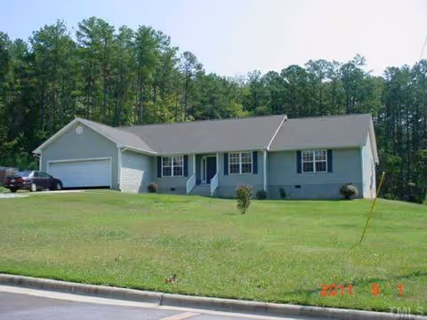 Single-story light-blue care home with a two-car garage, grassy front yard, and trees behind.