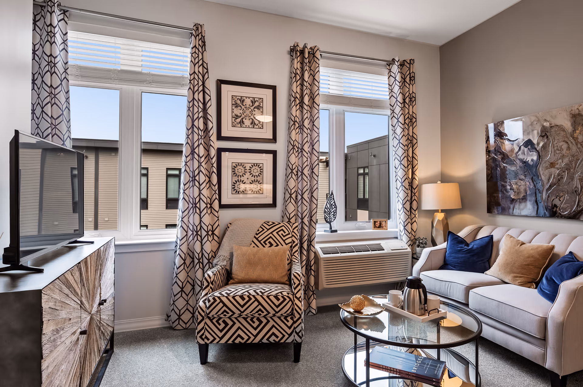 A cozy living room with a patterned armchair and a beige sofa adorned with blue and tan pillows. There are two large windows with geometric patterned curtains letting in natural light. A glass coffee table holds a tray with a coffee pot and cups, and a wooden TV stand supports a flat-screen television. Wall art and a table lamp add decorative touches.