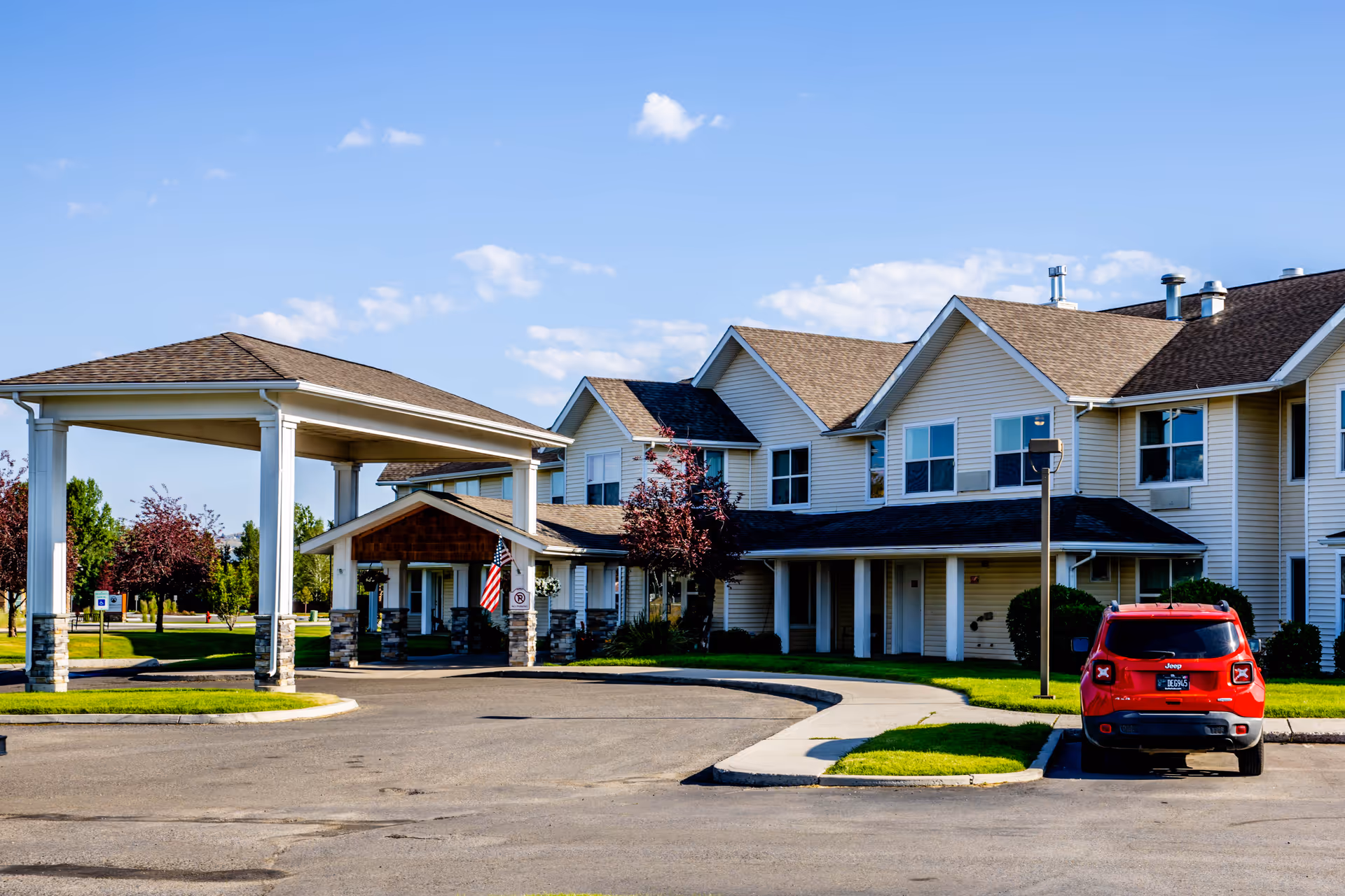 Exterior view of The Springs at Butte senior living facility showing a two-story building with beige siding and brown roofs, a covered entrance with white pillars, an American flag, a red Jeep parked in front, and a clear blue sky with a few clouds.