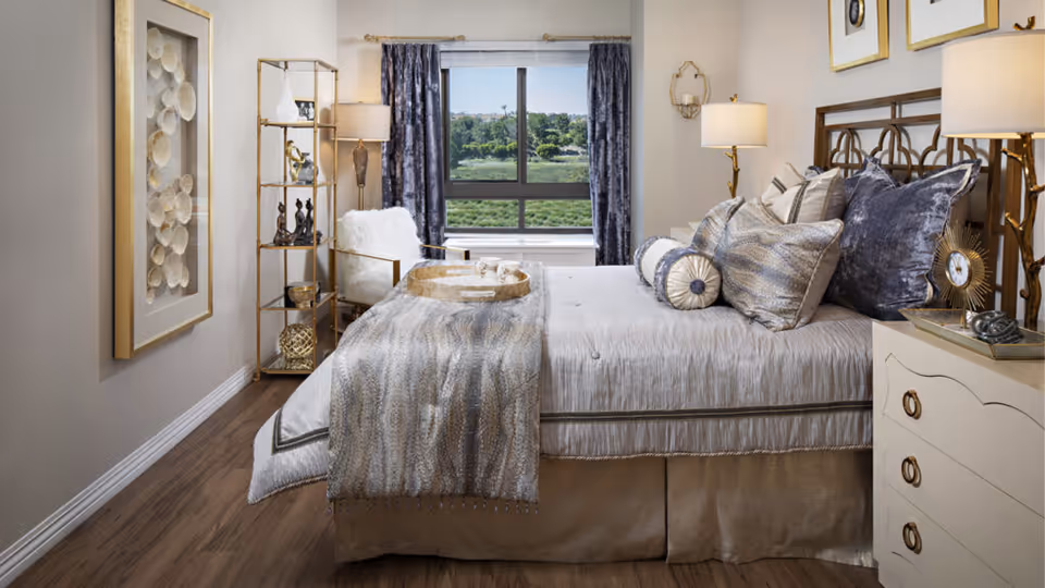 A well-decorated bedroom with a large bed adorned with multiple pillows and a textured throw blanket. There is a window with dark curtains showing a green outdoor view. The room features a white nightstand with a decorative clock and lamp, a gold metal shelving unit with decorative items, a white armchair with a fur throw, and framed wall art. The floor is wooden.
