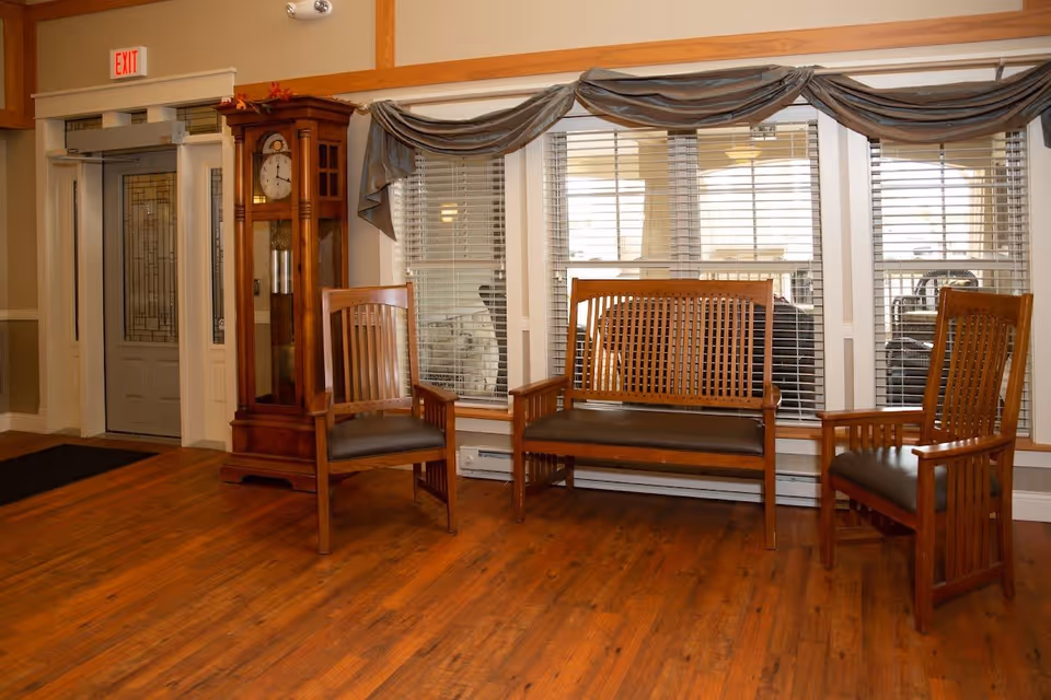 Lobby seating area with wooden chairs and a bench, a grandfather clock, and large windows beside an entrance door.
