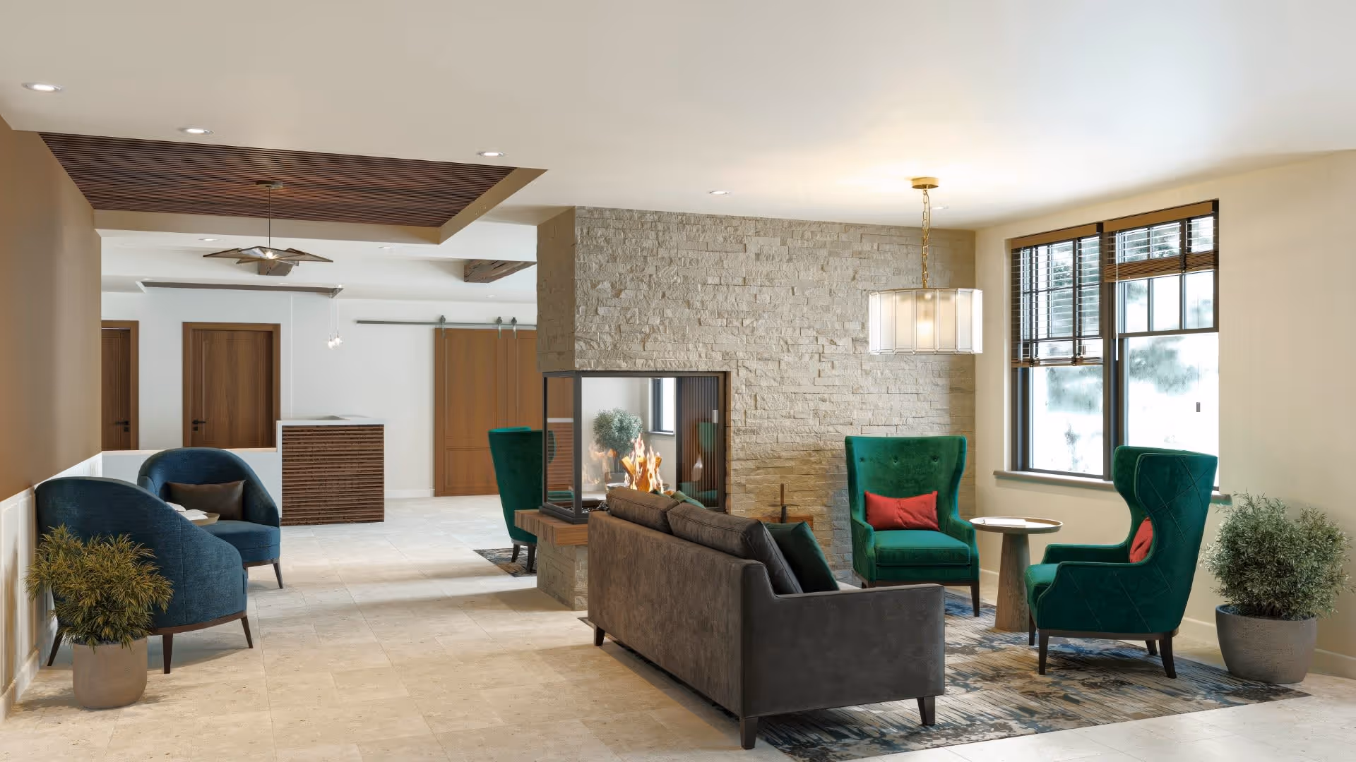 A modern living room area featuring a central three-sided glass fireplace with a stone surround. The room includes a dark gray sofa, two green wingback chairs with red cushions, a small round wooden table, and two blue armchairs with a small table between them. There are large windows with wooden blinds, potted plants, and a ceiling light fixture above the seating area. The flooring is light-colored tile, and the ceiling has recessed lighting and a wooden accent panel.