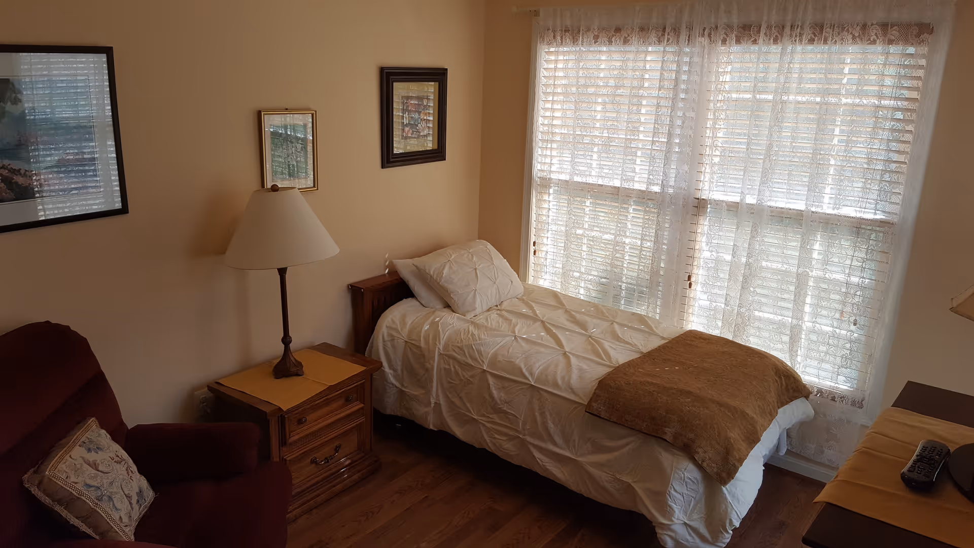 A cozy bedroom with a single bed covered in white bedding and a brown throw blanket. Next to the bed is a wooden nightstand with a lamp. There is a maroon armchair with a decorative pillow, and three framed pictures hang on the beige wall. A large window with sheer white curtains allows natural light to fill the room.