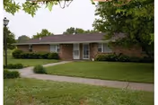 Single-story brick building with a dark shingled roof, surrounded by green grass and trees, with a concrete walkway leading to the front door.