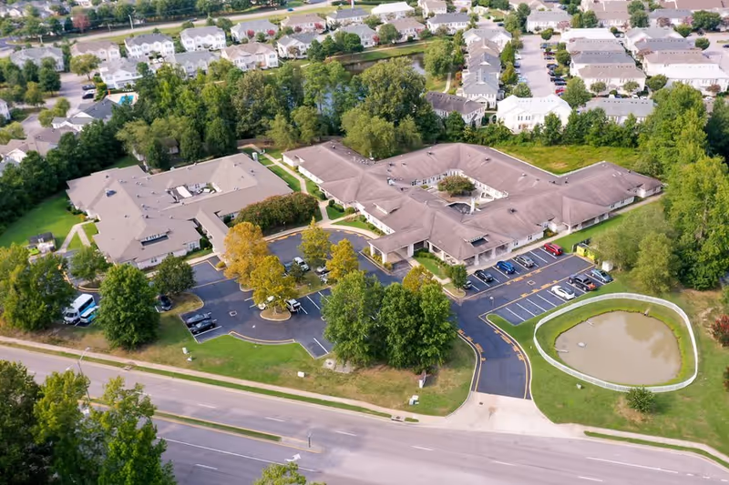 Aerial view of Chesapeake Place Senior Living facility showing a large building complex with multiple wings, surrounded by trees, parking lots with cars, a small pond with a white fence, and nearby residential houses.