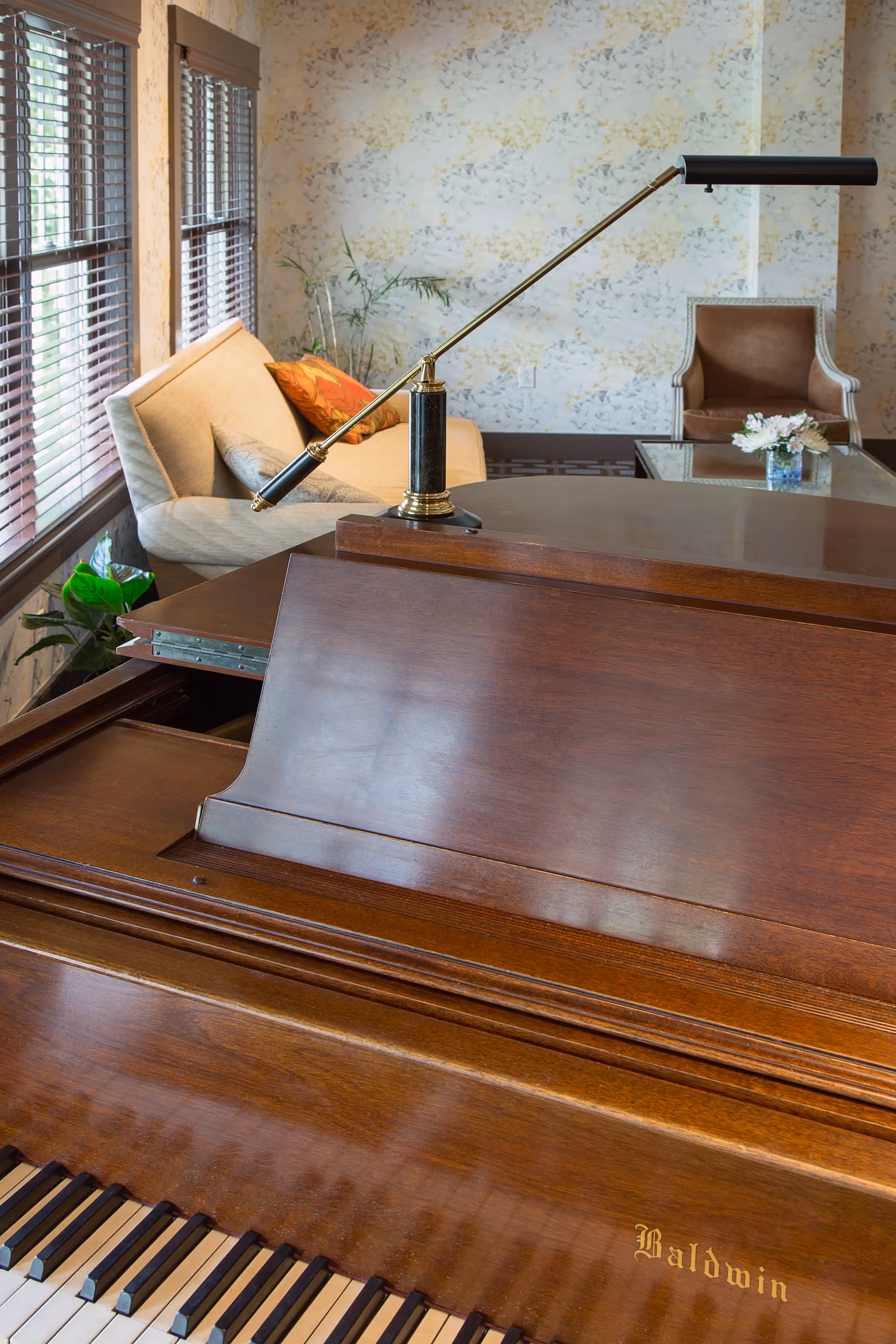 Interior view of a cozy living room area featuring a wooden Baldwin grand piano in the foreground with a piano lamp attached. In the background, there is a beige sofa with orange and beige cushions, a glass coffee table with a small flower arrangement, a brown armchair, and a wall with floral wallpaper. Large windows with wooden blinds allow natural light to fill the room.