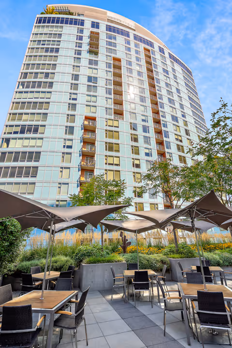 Curved glass high-rise building overlooking an outdoor patio with tables, chairs, umbrellas, and landscaping.