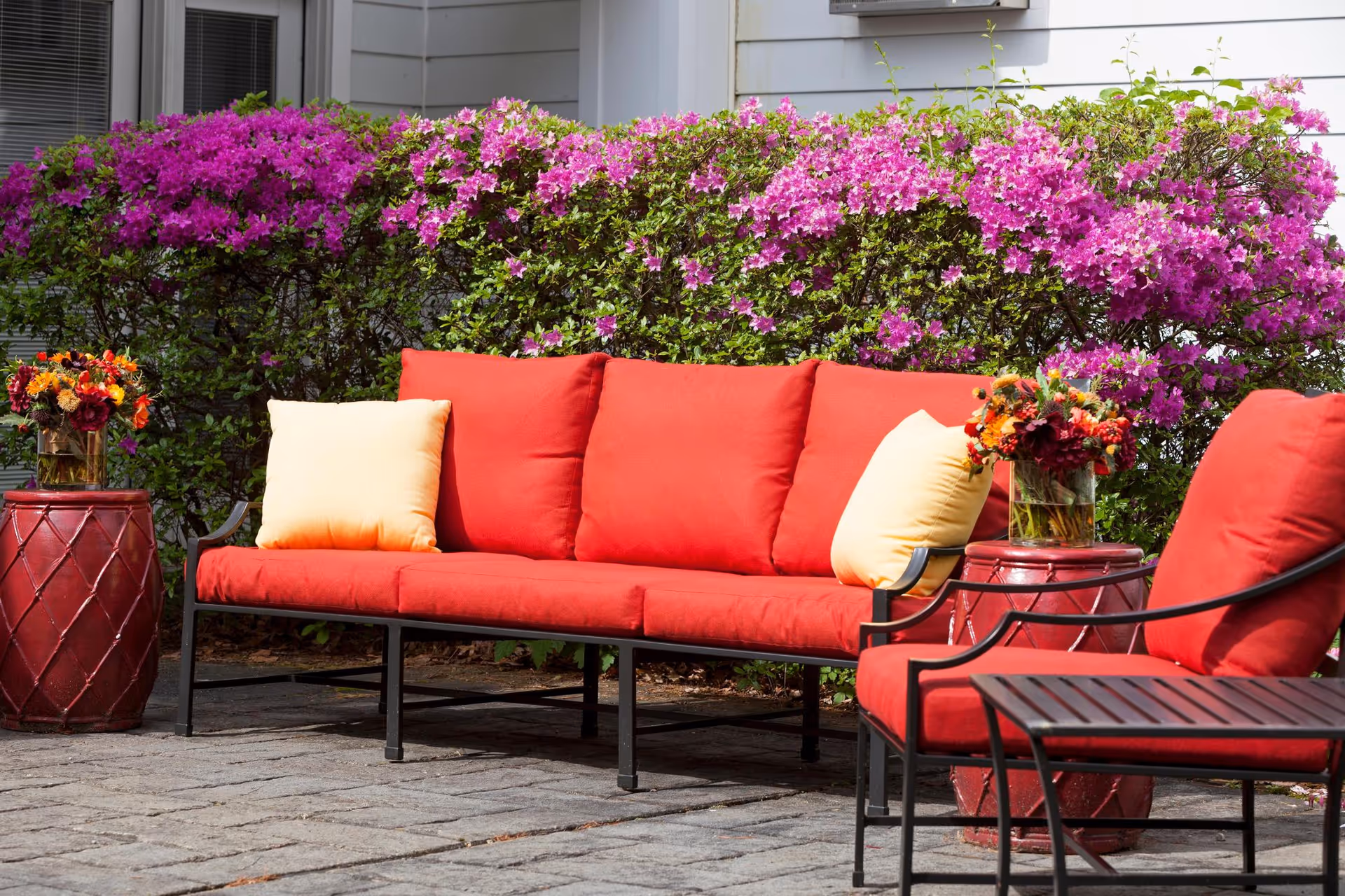 Red-cushioned outdoor sofa and chairs with yellow pillows on a patio in front of blooming purple azalea bushes.