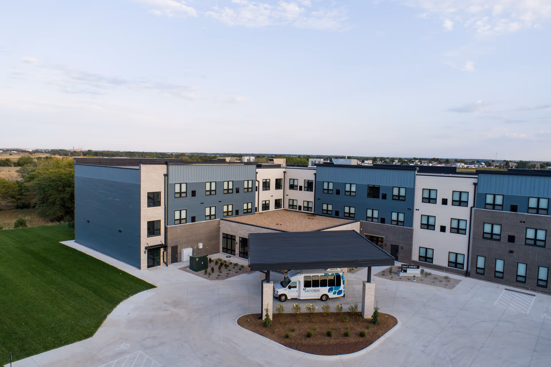 Exterior view of Salt Creek Senior Living facility showing a modern three-story building with a covered entrance where a Salt Creek shuttle bus is parked. The building features a mix of blue, white, and brick facade with multiple windows. Surrounding the building is a paved driveway and landscaped greenery.
