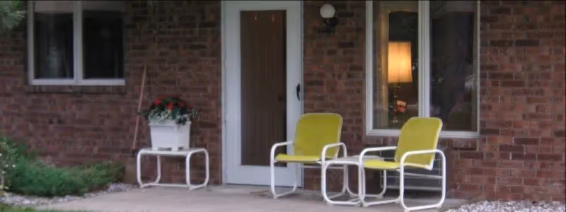 Outdoor patio area with two yellow metal chairs and a white metal table holding a white planter with red flowers, set against a brick wall with a door and two windows.