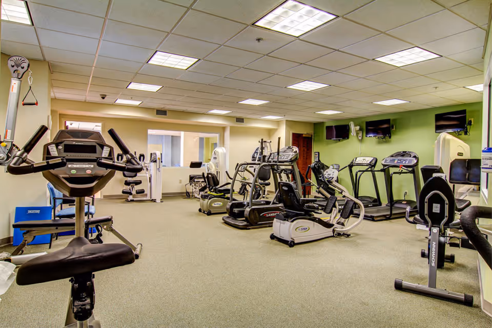 Interior view of a fitness room with various exercise equipment including stationary bikes, treadmills, and elliptical machines. The room has a green accent wall with three mounted flat-screen TVs and a beige carpeted floor under fluorescent ceiling lights.