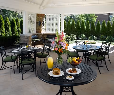 Covered outdoor patio with wrought-iron dining tables and chairs set with dishes, a vase of flowers, pancakes, juice, and fruit.