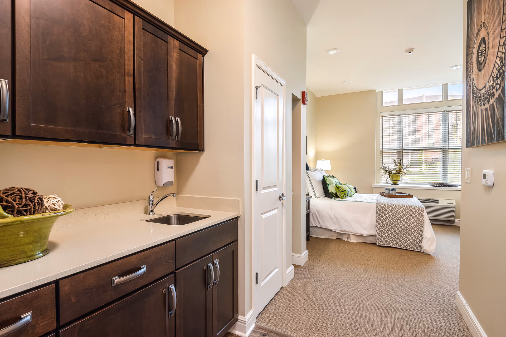 Interior view of a senior living facility room showing a kitchenette with dark wood cabinets and a small sink on the left. The room extends to a bedroom area with a neatly made bed, decorative pillows, a bedside lamp, and a window with blinds letting in natural light. A piece of wall art is visible on the right wall.