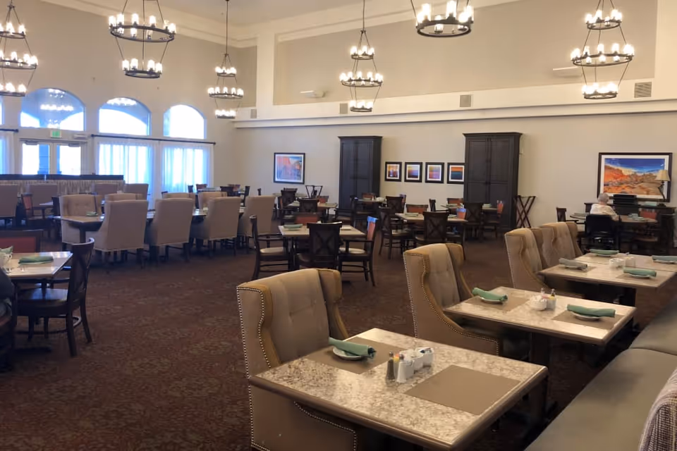 A spacious dining room with multiple tables and chairs arranged neatly. The tables are set with placemats, napkins, and condiments. The room features high ceilings with several chandeliers, large windows with curtains, and framed artwork on the walls. The overall ambiance is warm and inviting.