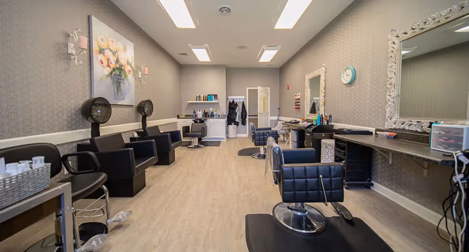 Interior view of a salon area in a senior living facility with multiple black salon chairs, hair dryers, mirrors with ornate white frames, and shelves with hair care products. The room has light wood flooring, patterned wallpaper, and bright overhead lighting.