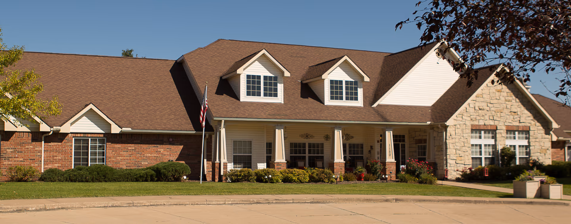 Single-story brick and stone senior living building front with a covered porch, dormer windows, an American flag, and landscaped lawn.