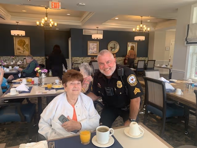 An elderly woman sitting at a dining table with a cup of coffee and a glass of orange juice, smiling with a police officer who is kneeling beside her. Other elderly people are seated at tables in the background in a well-lit dining room with chandeliers and framed artwork on the walls.