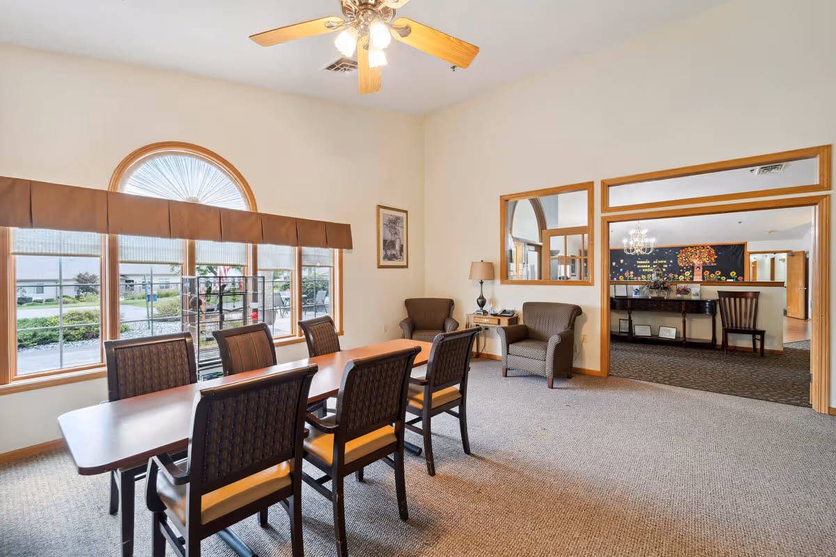 A well-lit room with a rectangular wooden table surrounded by six chairs. Two armchairs and a small side table with a lamp are positioned near a wall with a framed picture. Large windows with a half-circle window above provide natural light and a view of the outside. An open doorway with wooden trim leads to another room with a piano, chairs, and a decorative wall display.