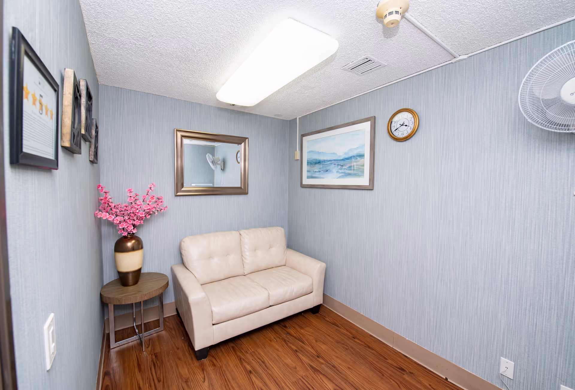 Small sitting room with a cream loveseat, side table with pink flowers, a wall mirror, artwork and a clock.