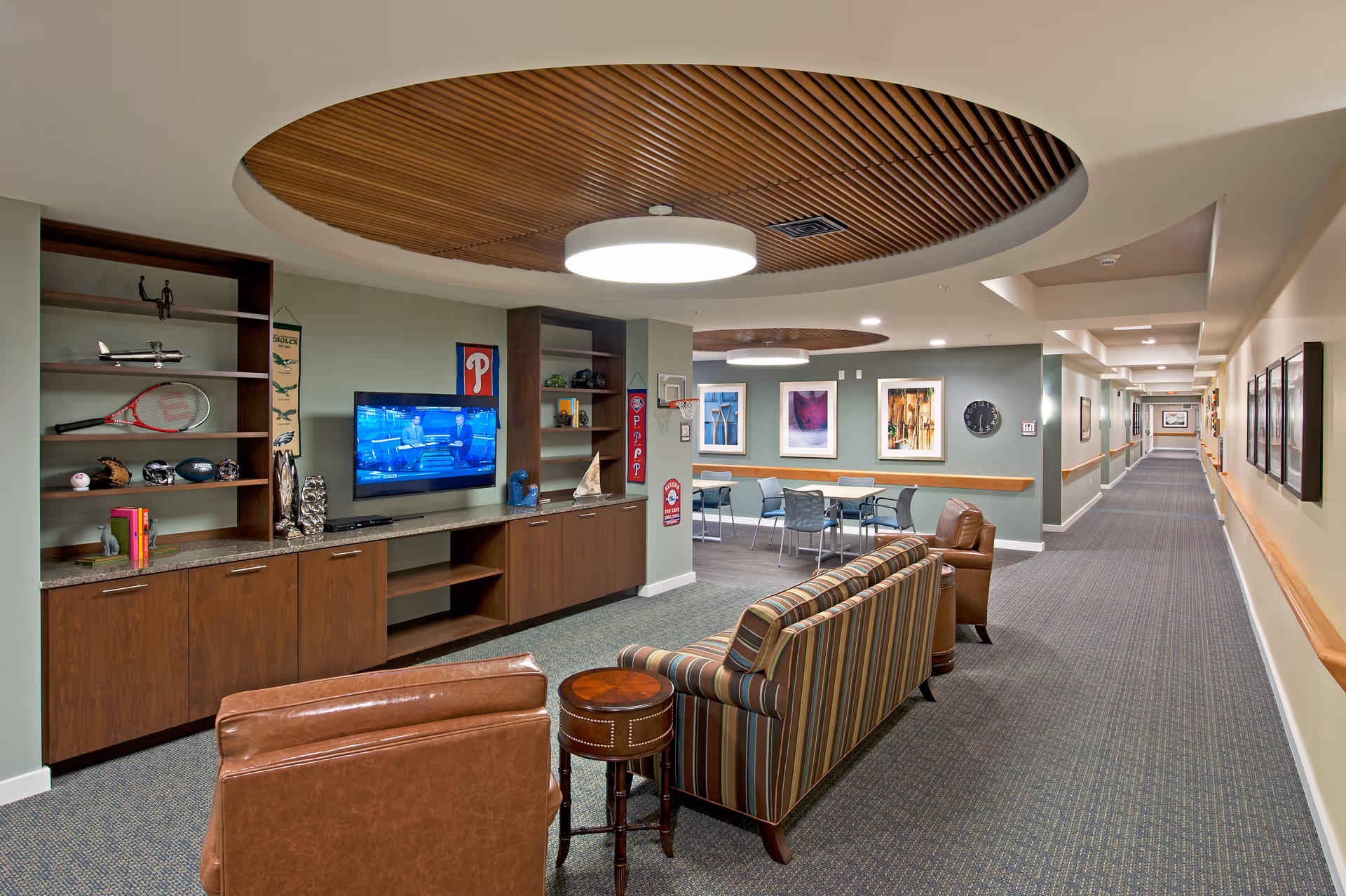 A senior living facility common area with a long hallway on the right and a seating area on the left. The seating area includes a striped sofa, two brown leather chairs, and a small round wooden table. Behind the seating area is a built-in wooden cabinet with shelves displaying sports memorabilia and a flat-screen TV mounted on the wall. The ceiling features circular wooden paneling with a large round light fixture. Further back, there is a dining or activity area with tables and chairs, and framed artwork on the walls.