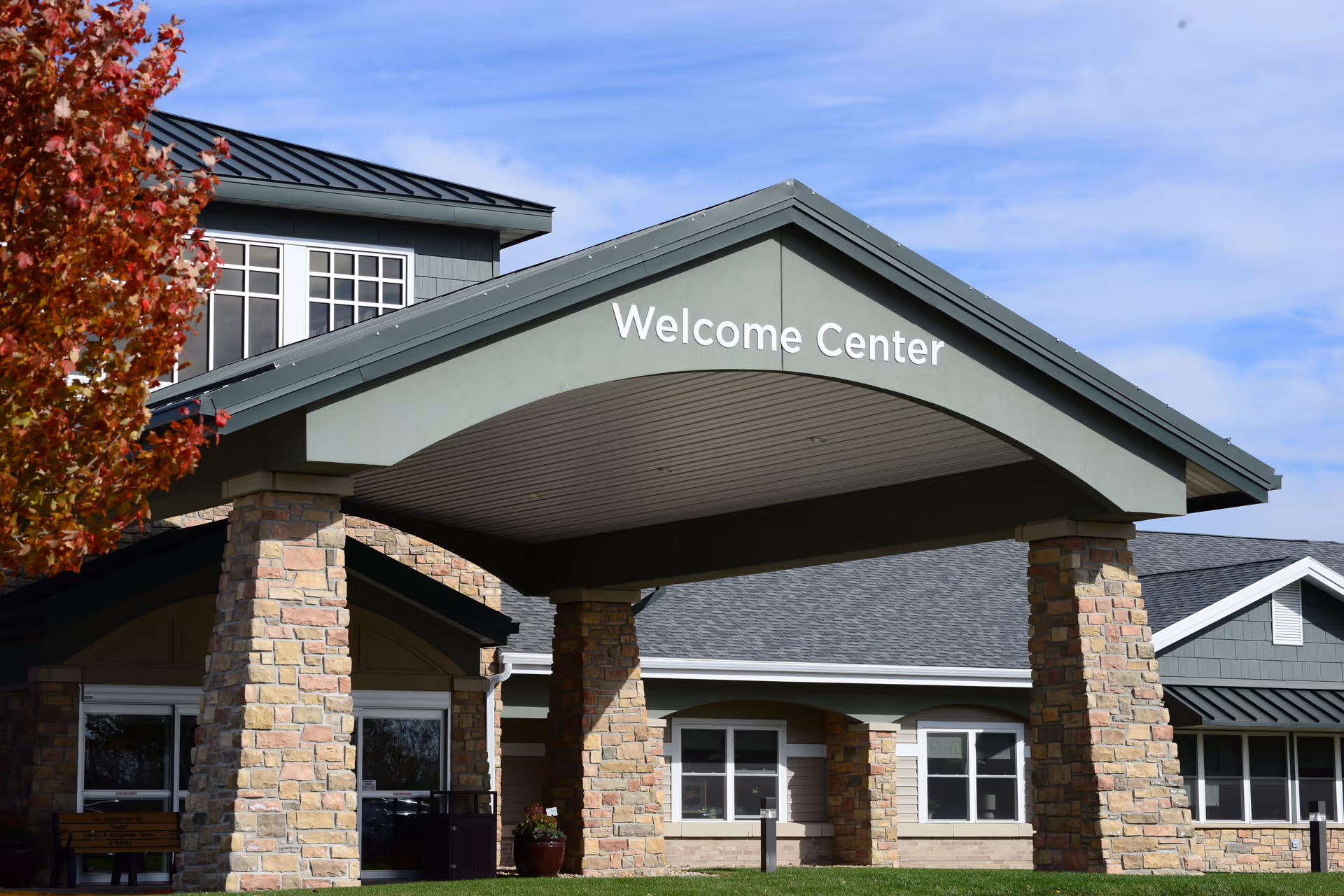 Exterior view of a building with a covered entrance supported by stone pillars. The entrance has a sign that reads 'Welcome Center'. The building has multiple windows and a dark roof. A tree with red autumn leaves is visible on the left side under a blue sky with some clouds.