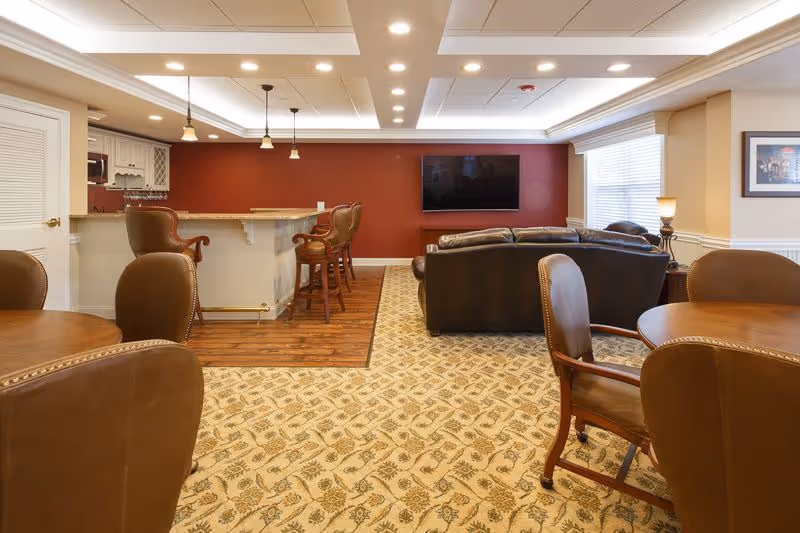 Well-lit communal lounge with a bar, high stools, leather sofas, round tables, and a wall-mounted TV.