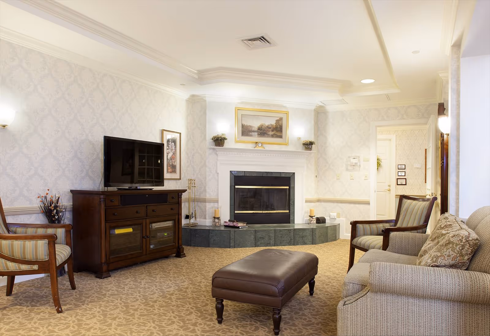 A cozy living room with patterned wallpaper and carpet, featuring a fireplace with a framed painting above it. There is a wooden TV stand with a flat-screen TV, two armchairs with striped cushions, a beige sofa with a decorative pillow, and a brown leather ottoman in the center.
