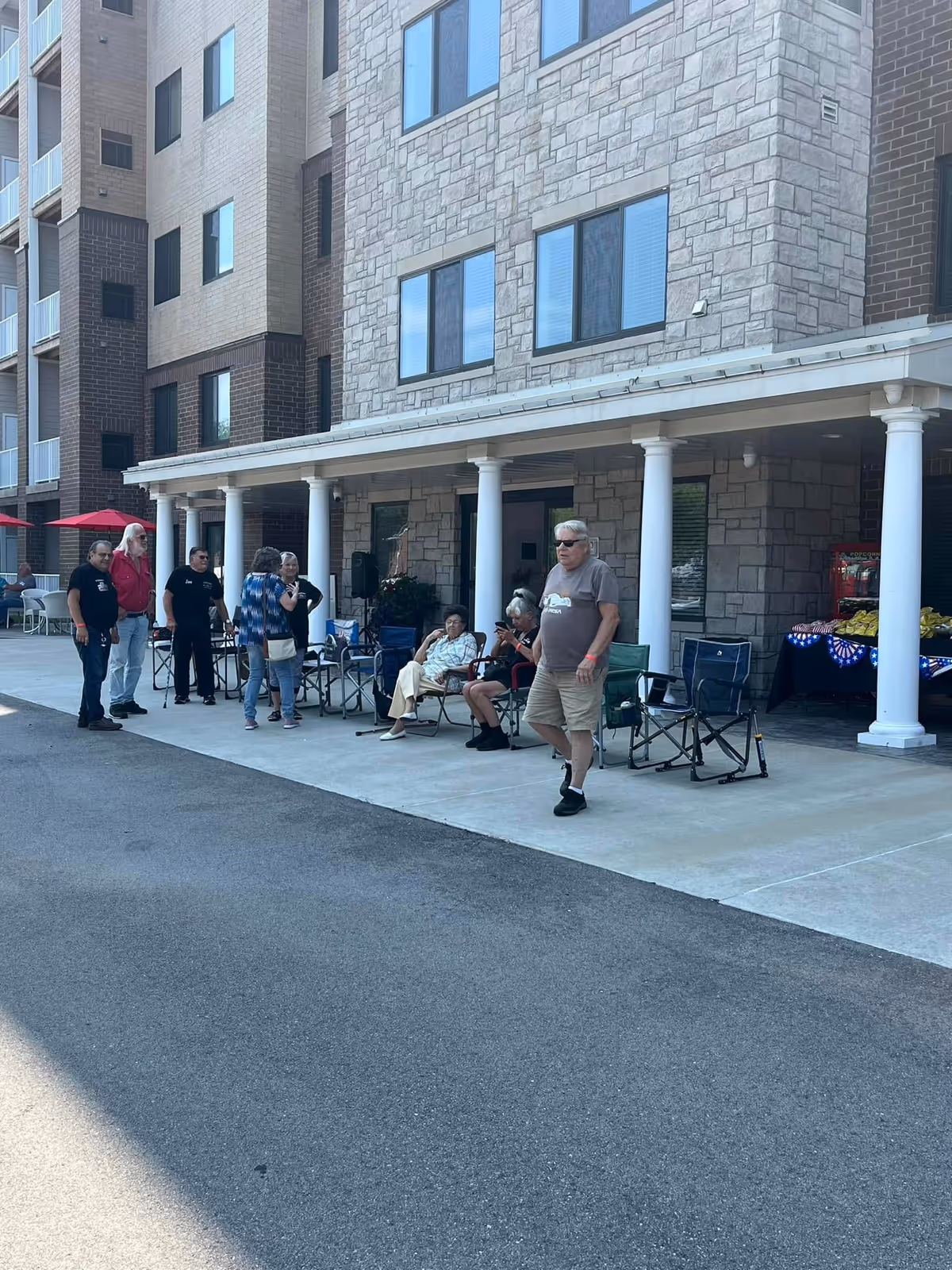 Group of older adults sitting and standing under the covered entrance of a multi-story stone-and-brick building with white columns.