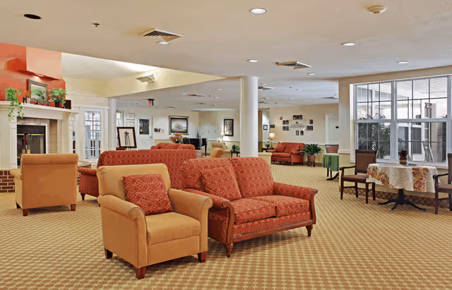 A spacious senior living facility common area with patterned carpet, several upholstered armchairs and sofas in warm tones, a fireplace with plants on the mantle, framed artwork on the walls, and a table with chairs near large windows letting in natural light.