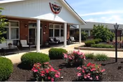 Front entrance of a single-story brick health care building with a covered porch and outdoor seating, surrounded by manicured shrubs and pink flowering plants.
