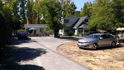 A gray, chalet-style senior living building sits behind a curved driveway with cars and trees.