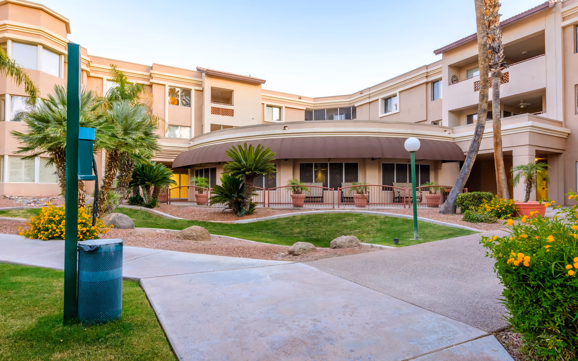 Exterior view of a senior living facility courtyard with a curved building facade, palm trees, green grass, flowering bushes, and paved walkways under a clear sky.