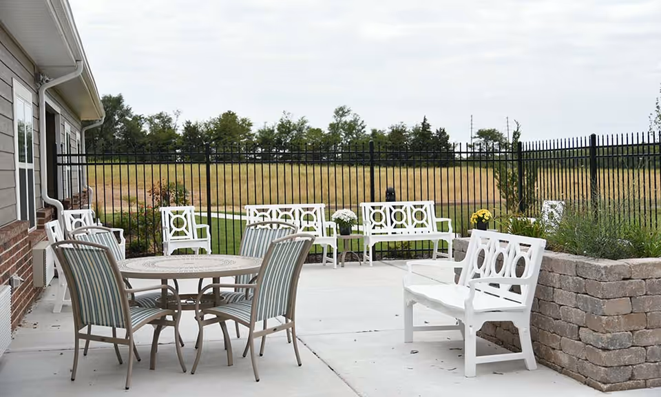 Outdoor patio area with white benches and a round table with four striped cushioned chairs. The patio is adjacent to a building with brick and siding exterior, surrounded by a black metal fence and a grassy field beyond.