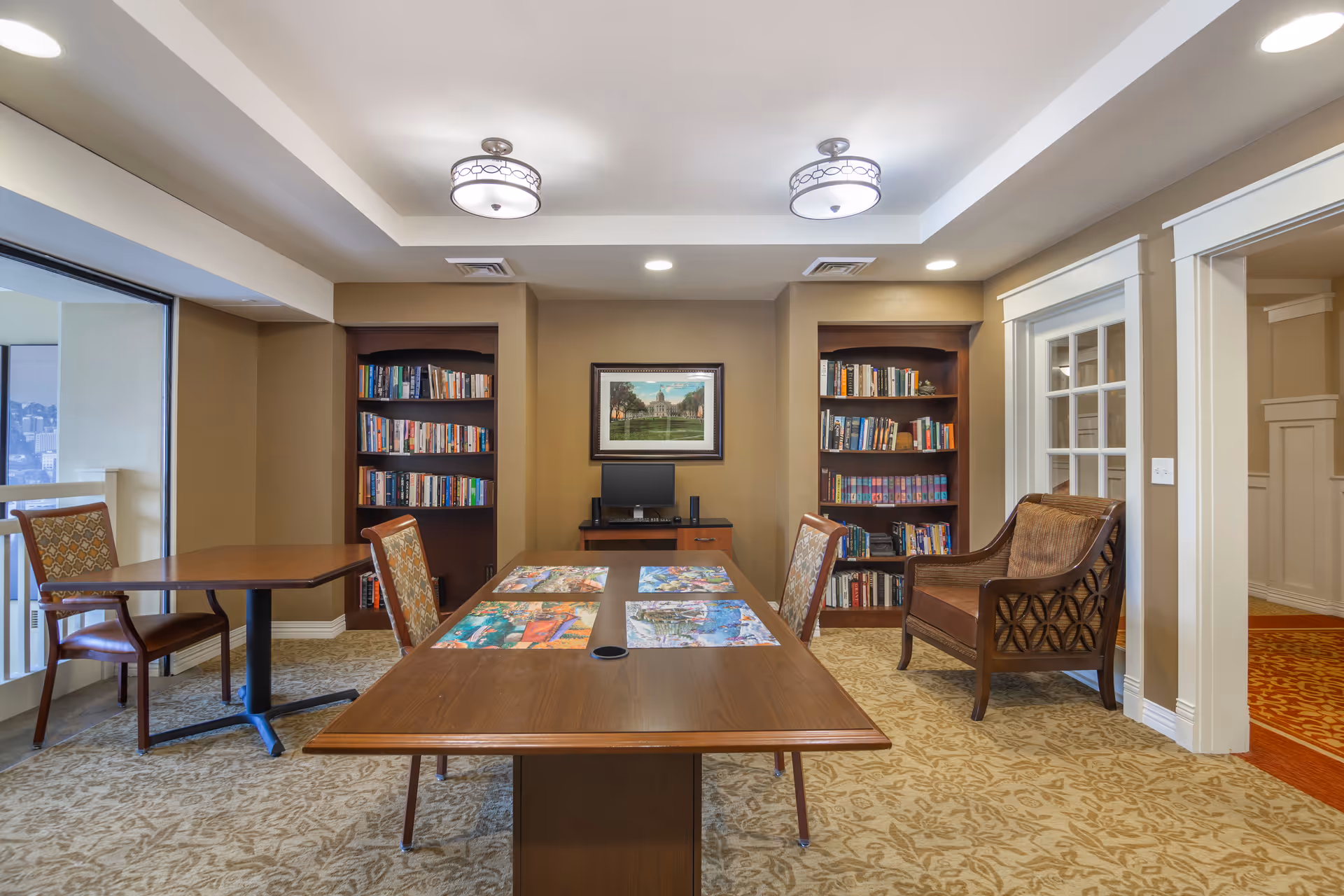 A well-lit interior room with a large wooden table in the center, surrounded by chairs. The table has colorful puzzles or artwork on it. Behind the table are two built-in bookshelves filled with books and a small desk with a computer monitor. The room has beige walls, patterned carpet, and two ceiling light fixtures. There is a large window on the left side and a doorway on the right.