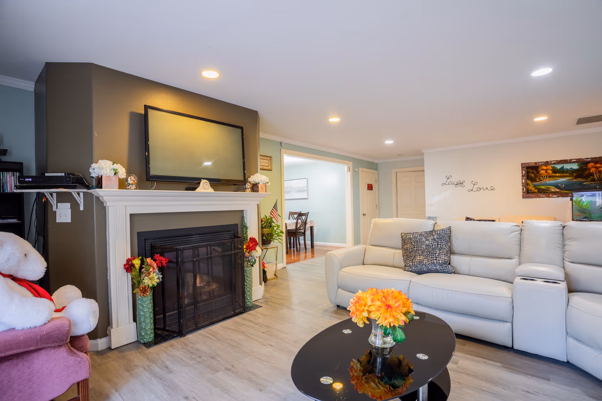 A cozy living room with a white sectional sofa adorned with a patterned pillow, a black oval coffee table with a vase of orange flowers, a fireplace with a TV mounted above it, and a pink armchair with a large white teddy bear sitting on it. The walls are light-colored with a decorative sign that says 'Laugh Love' and a framed painting. The room is well-lit with recessed ceiling lights and has a view into an adjacent dining area.