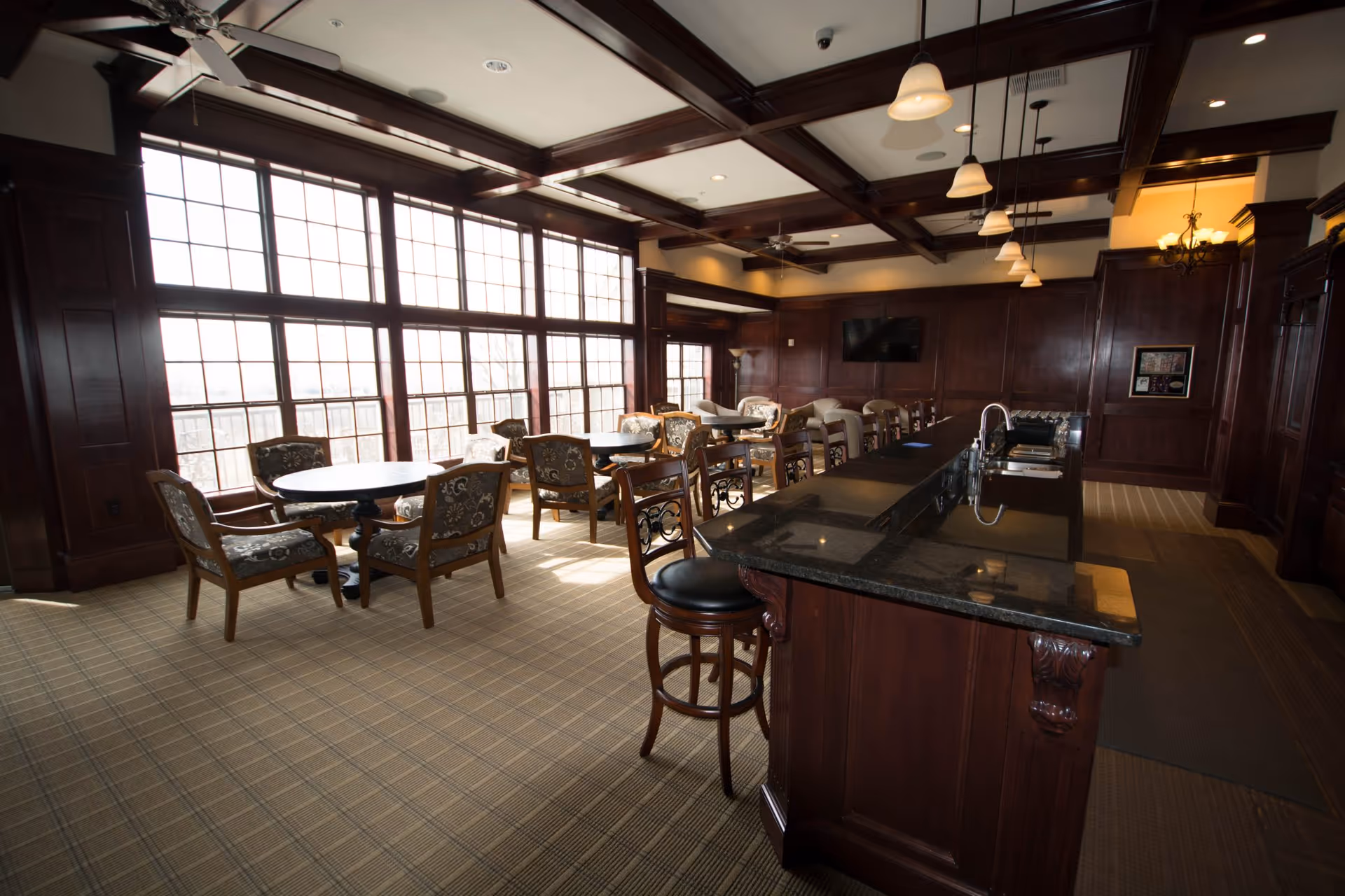 A spacious, elegant common area with large windows letting in natural light. The room features dark wood paneling and ceiling beams, a long granite countertop with bar stools, several round tables with upholstered chairs, and a wall-mounted TV. Pendant lights hang from the ceiling, and the carpet has a subtle plaid pattern.